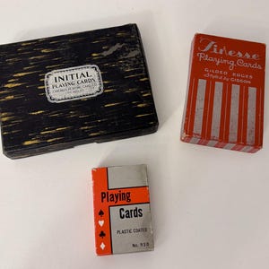 May include: Three boxes of playing cards. The first box is black with gold lettering and says "Initial Playing Cards". The second box is red and white with gold lettering and says "Finesse Playing Cards Gilded Edges  Stuka by CISSON". The third box is white with black lettering and says "Playing Cards Plastic Coated No. 930".
