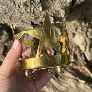 Puede incluir: Un portavelas de lat&oacute;n con dise&ntilde;o de estrella. El soporte tiene forma cil&iacute;ndrica con formas de estrella recortadas y est&aacute; sostenido por tres peque&ntilde;os pies redondos. El lat&oacute;n tiene un tono dorado c&aacute;lido.