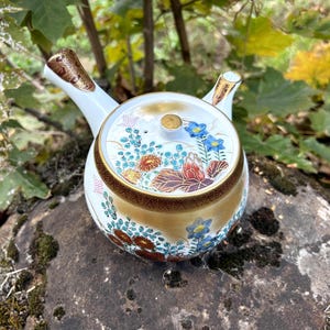 Puede incluir: Una tetera kyusu de cerámica blanca con asa lateral y tapa decorada con intrincados motivos florales en azul, rojo y oro. El cuerpo de la tetera presenta un degradado dorado con detalladas ilustraciones botánicas y bordes en color oro.