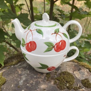 May include: White ceramic teapot and teacup set with a cherry design. The teapot has a lid and handle, and the teacup fits snugly underneath. The cherries are red with green leaves, and the set is on a mossy rock.