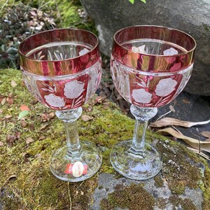 Pode incluir: Duas taças de vinho de cristal com borda vermelha e design floral gravado. Os óculos têm uma haste e uma base transparentes. Os óculos estão em uma rocha coberta de musgo.