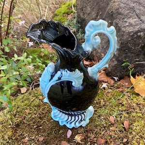 May include: A black ceramic pitcher with a blue and white floral design. The pitcher has a wide mouth and a handle that is shaped like a scroll. The pitcher is sitting on a bed of moss.