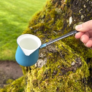 Könnte beinhalten: Eine kleine blaue Emaille-Kaffeekanne mit weißem Innenraum und langem, schwarzem Griff. Die Kanne ist mit einer klaren Flüssigkeit gefüllt. Der Hintergrund zeigt einen moosbewachsenen Baumstamm und grünes Gras.