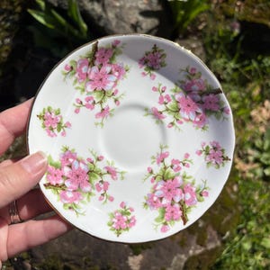 K&ouml;nnte beinhalten: Wei&szlig;e Keramikuntertasse mit goldenem Rand, verziert mit rosa Bl&uuml;tenclustern und gr&uuml;nen Bl&auml;ttern. Die Untertasse hat eine zentrale Vertiefung f&uuml;r eine Teetasse. Ein zartes und elegantes Design.