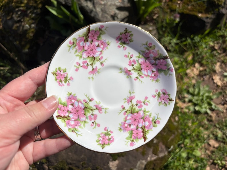 K&ouml;nnte beinhalten: Wei&szlig;e Keramikuntertasse mit goldenem Rand, verziert mit rosa Bl&uuml;tenclustern und gr&uuml;nen Bl&auml;ttern. Die Untertasse hat eine zentrale Vertiefung f&uuml;r eine Teetasse. Ein zartes und elegantes Design.