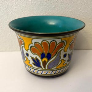 May include: A ceramic planter with a flared rim and a teal interior. The exterior features a white background decorated with a repeating pattern of orange, blue, and purple floral motifs, accented by yellow shapes and a thin geometric border.