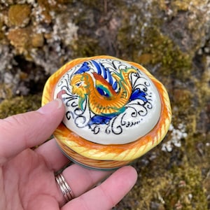 May include: A small, round ceramic trinket box with a lid. The lid features a colorful design of a mythical creature in orange, yellow, blue, and green. The box has a yellow and orange braided border. The box is held in a hand.