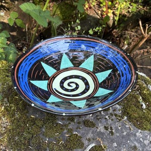 Könnte beinhalten: Eine handbemalte Keramikschale mit einem Sonnenmuster. Die Schale hat eine schwarze Außenseite mit einem blauen und orangefarbenen Rand. Das Innere zeigt ein türkisfarbenes Sonnenmuster mit einem weißen Spiralmittelpunkt. Die Schale hat einen Durchmesser von ca. 20 cm.