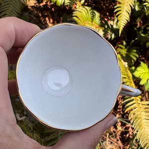 May include: A white teacup with a gold rim and handle, viewed from above. The cup is held in a hand, with green foliage in the background. The cup has a simple, elegant design.