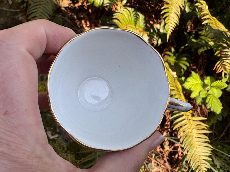 May include: A white teacup with a gold rim and handle, viewed from above. The cup is held in a hand, with green foliage in the background. The cup has a simple, elegant design.
