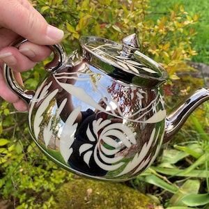 May include: A silver teapot with a white floral design. The teapot has a shiny, reflective surface and a lid with a pointed knob.