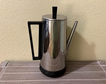 Op de afbeelding: Een hoge, cilindrische koffiepercolator met een gepolijste chromen afwerking. Het apparaat heeft een zwart, hoekig handvat, een bijpassende zwarte dekselknop en een zwarte voet. Een lange, slanke, schuine tuit steekt uit de metalen behuizing.