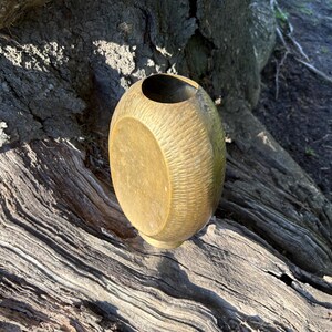 Peut inclure: Un vase ovale de couleur dorée avec une surface texturée. Le vase a un côté lisse et plat et une ouverture circulaire en haut. Le vase repose sur une surface en bois vieilli et texturé.