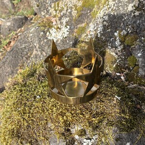 Puede incluir: Un portavelas de metal dorado con recortes de estrellas. El portavelas es circular con una base plana y un borde. Los recortes de estrellas est&aacute;n espaciados uniformemente alrededor de la parte superior. El portavelas est&aacute; sobre una roca cubierta de musgo.