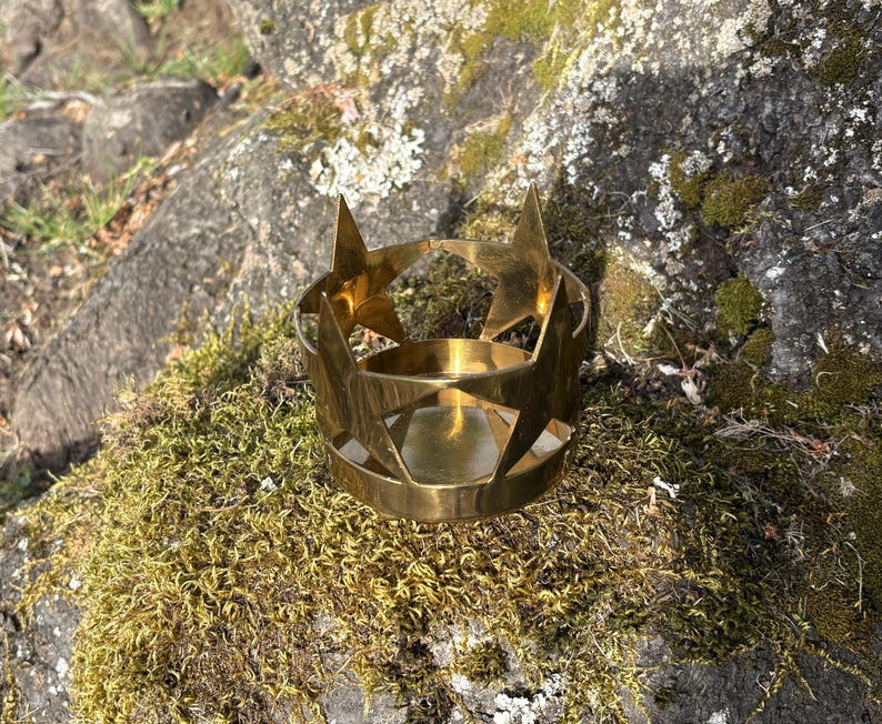 Puede incluir: Un portavelas de metal dorado con recortes de estrellas. El portavelas es circular con una base plana y un borde. Los recortes de estrellas est&aacute;n espaciados uniformemente alrededor de la parte superior. El portavelas est&aacute; sobre una roca cubierta de musgo.