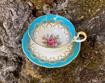 May include: A vintage teacup and saucer set with a turquoise blue rim and gold trim. The cup features a floral design with a pink rose and white background.