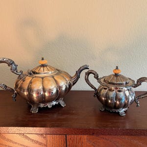 May include: A silver-colored tea set, including a teapot and a sugar bowl, sits on a wooden surface. The teapot has a curved handle and a pumpkin-shaped body. The sugar bowl has a similar design. Both have orange-colored finials.