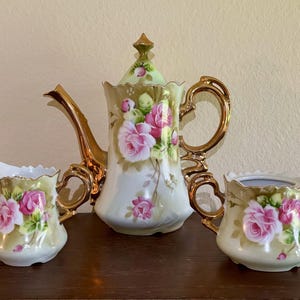 May include: A vintage tea set comprising a teapot, creamer, and sugar bowl. The set is adorned with pink roses and gold detailing. The teapot has an elaborate design with a lid and a curved spout. The creamer and sugar bowl feature matching floral patterns.