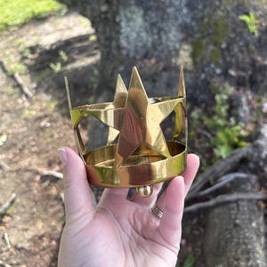 Puede incluir: Un objeto de metal dorado en forma de corona con recortes de estrellas y un dise&ntilde;o de estrella central. La corona tiene una base redondeada y puntas. El objeto se sostiene en una mano sobre un fondo exterior borroso.