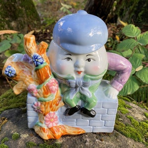 Könnte beinhalten: Eine Keramik-Teekanne in Form der Figur Humpty Dumpty aus dem Kinderreim. Die Teekanne ist in leuchtenden Farben bemalt, darunter Blau, Grün, Rosa und Orange. Die Teekanne steht auf einer Mauer aus Ziegeln, aus der ein kleiner Baum wächst.