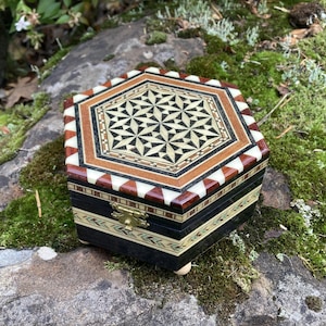 May include: A decorative wooden box with a geometric design on the lid. The box features inlaid wood in shades of brown, black, and cream. The box is hexagonal and rests on small, rounded feet. The clasp is gold-colored.