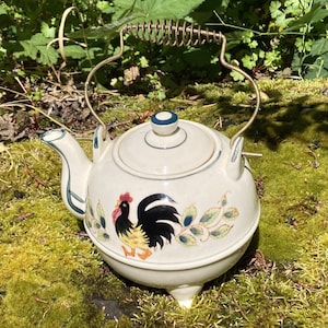 Könnte beinhalten: Cremefarbene Keramik-Teekanne mit Hahnmotiv. Die Teekanne hat einen runden Körper, einen gebogenen Ausguss und einen Deckel mit blauem Akzent. Sie verfügt über einen Metallgriff mit einer spiralförmigen Oberseite. Der Hahn ist in Schwarz, Gelb und Rot dargestellt.