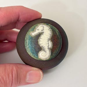 May include: A round, dark brown ceramic object featuring a central circular inlay. The inlay displays a white seahorse relief against a background of iridescent, shimmering green, blue, and copper tones that catch the light.