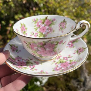 K&ouml;nnte beinhalten: Ein wei&szlig;es Tee-Set mit einem floralen Muster in Rosa und Gr&uuml;n. Die Tasse und Untertasse haben einen goldenen Rand. Die Teetasse wird in einer Hand vor einem verschwommenen gr&uuml;nen Hintergrund gehalten.