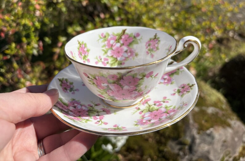 K&ouml;nnte beinhalten: Ein wei&szlig;es Tee-Set mit einem floralen Muster in Rosa und Gr&uuml;n. Die Tasse und Untertasse haben einen goldenen Rand. Die Teetasse wird in einer Hand vor einem verschwommenen gr&uuml;nen Hintergrund gehalten.