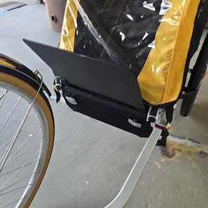 May include: A black and yellow bicycle trailer with a clear plastic window. The trailer is attached to a bicycle wheel with a tan tire. The trailer has a black storage compartment and a white metal arm.