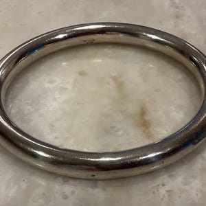 May include: A silver-colored, thick bangle bracelet. The bracelet is a solid, rounded band with a smooth, reflective surface. The word "MEXICO" is stamped on the inside of the bracelet. Jewelry.