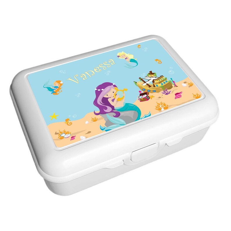 May include: White lunchbox with a colorful mermaid and a sunken ship design. The lunchbox has the name "Vanessa" printed on the front.
