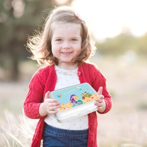 May include: A white lunchbox with a mermaid and a treasure chest design. The lunchbox has a blue background with gold accents. The name "Vanessa" is printed on the front of the lunchbox.