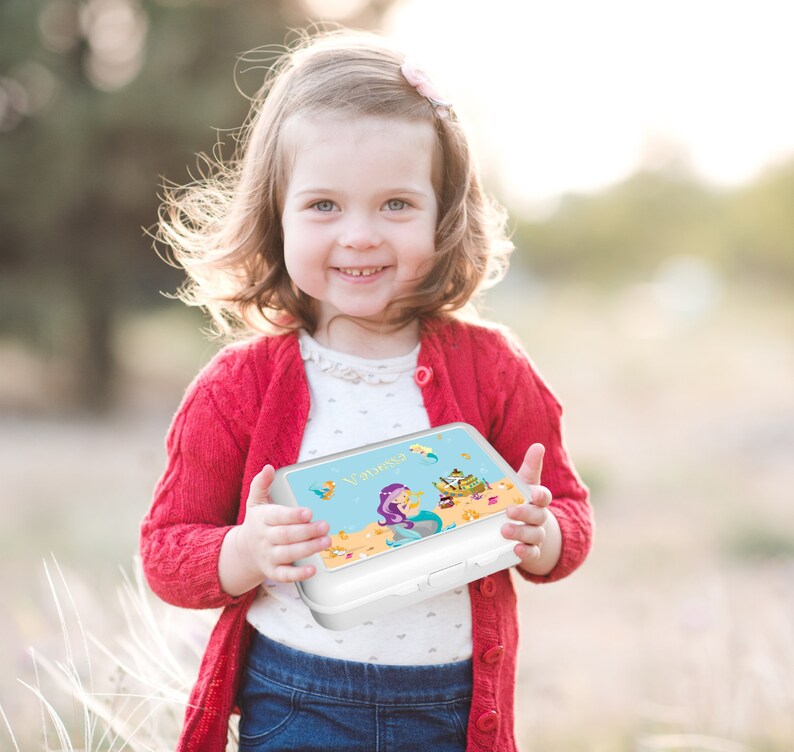 May include: A white lunchbox with a mermaid and a treasure chest design. The lunchbox has a blue background with gold accents. The name "Vanessa" is printed on the front of the lunchbox.