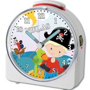 Könnte beinhalten: Wecker in Weiß mit einem Piraten-Motiv. Das Zifferblatt zeigt einen Cartoon-Piraten, einen Papagei, ein Piratenschiff und den Namen "NIKLAS". Der Wecker hat eine rote Oberseite und einen grünen Weckschalter.