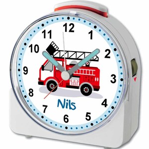 Könnte beinhalten: Weißer Wecker mit einem roten Feuerwehrauto-Motiv und dem Namen "Nils" auf dem Zifferblatt. Die Uhr hat blaue Zeiger und einen blauen und weißen Rand.