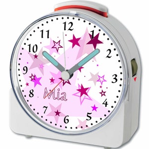 Könnte beinhalten: Weißer Wecker mit einem rosafarbenen Zifferblatt mit einem rosa-weißen Sternenmuster. Das Zifferblatt hat schwarze Zahlen und einen schwarzen Zeiger. Der Name "Mia" ist in Schwarz auf dem Zifferblatt geschrieben.
