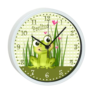 Könnte beinhalten: Eine weiße runde Uhr mit schwarzen Zahlen und Zeigern. Das Zifferblatt hat ein grün-weißes Karomuster mit grünem Gras und einem grünen Frosch mit rosa Herzen. Auf dem Zifferblatt steht auch der Name "Paulina".