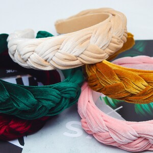 May include: Four braided velvet headbands in different colors: green, beige, mustard yellow, and pink. The headbands are stacked on top of each other.
