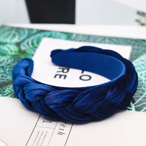 May include: A blue velvet headband with a braided design. The headband is on a white surface with a green leafy pattern.