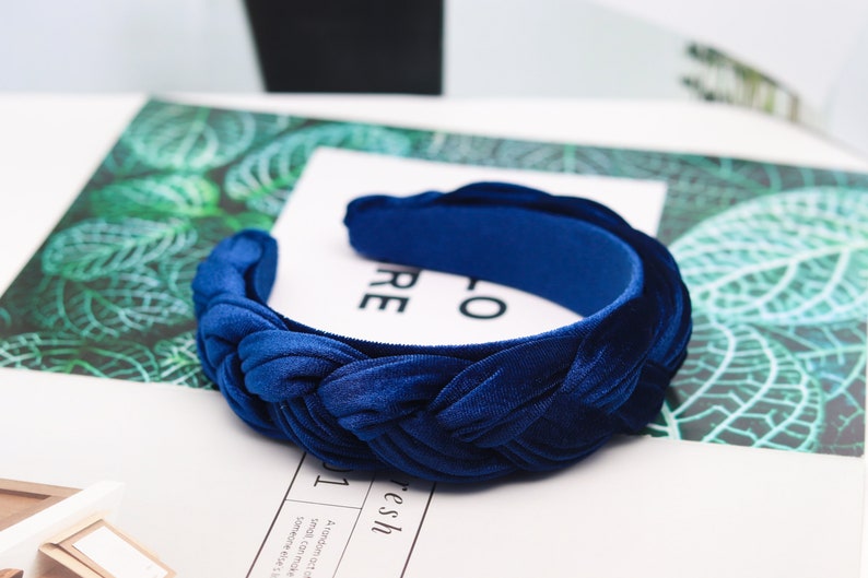 May include: A blue velvet headband with a braided design. The headband is on a white surface with a green leafy pattern.