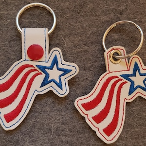 Może przedstawiać: Dwa białe skórzane breloki z haftowanymi gwiazdami i paskami w kolorze czerwonym, białym i niebieskim. Breloki mają srebrne kółka do kluczy.