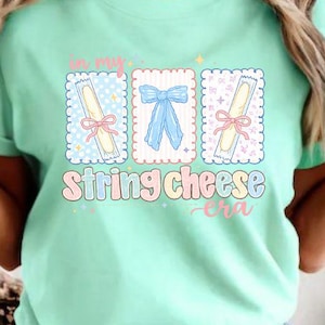 May include: A pale green t-shirt with the phrase "in my string cheese era." The design includes three illustrated rectangles with bow and string cheese images. The text and illustrations are in pastel shades.