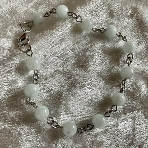May include: A delicate bracelet featuring round, white beads strung on a silver-colored chain. The bracelet has a heart-shaped clasp and is designed to be worn around the wrist. The beads are evenly spaced and create a simple, elegant look.