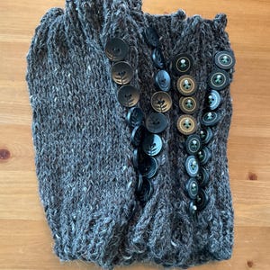 May include: A pair of dark gray, chunky knit leg warmers featuring a vertical row of decorative buttons on each side. The buttons are a mix of solid black and metallic gold-toned circular designs, each embossed with a small leaf or floral motif.