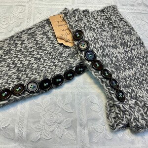 May include: Gray and white knit arm warmers with dark brown buttons. The arm warmers have a textured pattern and are adorned with a row of buttons along the edge. A small tag is attached to one of the arm warmers.