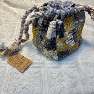 May include: A gray and yellow patchwork drawstring bag with a braided cotton cord handle. The bag is fully lined with two pockets and is hand stitched. The tag attached to the bag reads "100% Cotton Fully Lined 2 Pockets Slow Stitch Fully Hand Stitched Kantha Boro Machine wash and dry."