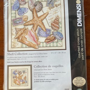 May include: A Dimensions counted cross stitch kit titled Shell Collection featuring a printed design of various seashells, a starfish, a blue butterfly, and vintage postcards. The package includes 14 count ivory Aida fabric measuring 15 by 15 inches.