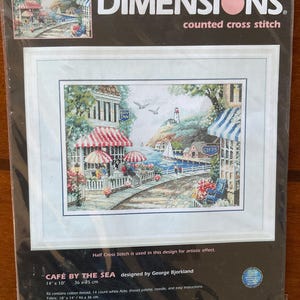 May include: A Dimensions counted cross stitch kit titled Café by the Sea, designed by George Bjorkland. The packaging displays a finished 14 inches by 10 inches coastal scene featuring a seaside cafe with red and white striped awnings.