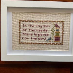 May include: A white framed cross-stitch embroidery on beige fabric featuring the text In the rhythm of the needle there is peace for the soul, accompanied by a small bird and a stack of colorful spools, all enclosed in a decorative border.